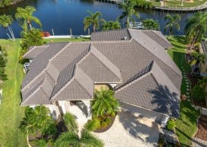 Aerial view of a large house with a tiled roof, surrounded by palm trees and landscaping, located beside a body of water with a dock.