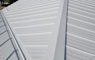 White metal roof with standing seam panels and a prominent ridge, viewed from above. A green utility truck and palm trees are visible in the background.