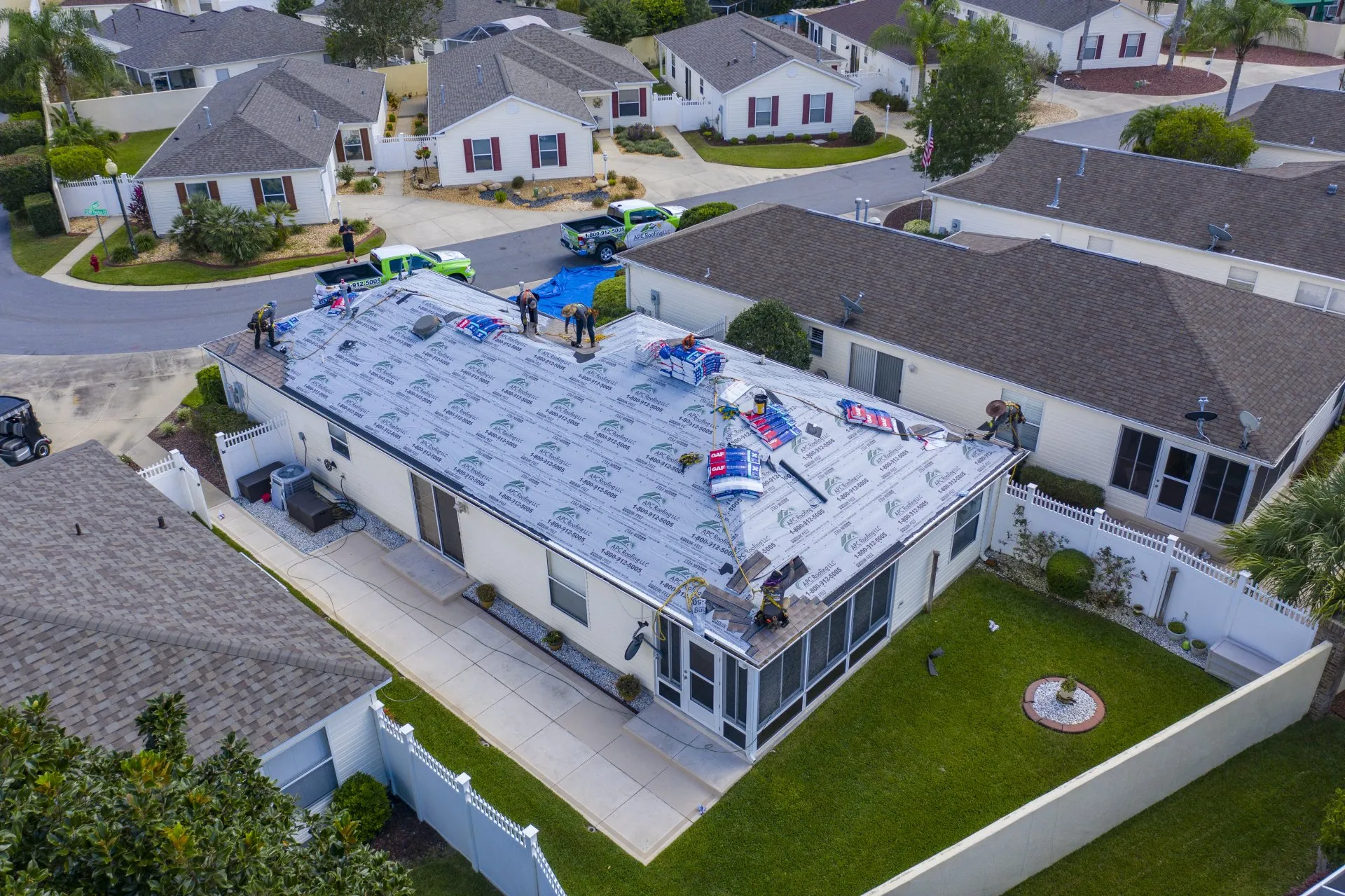 Aerial view of workers installing roofing materials on a single-story house in a residential neighborhood. Roofing supplies and tools are spread across the roof. Aerial view of workers installing roofing materials on a single-story house in a residential neighborhood. Roofing supplies and tools are spread across the roof.