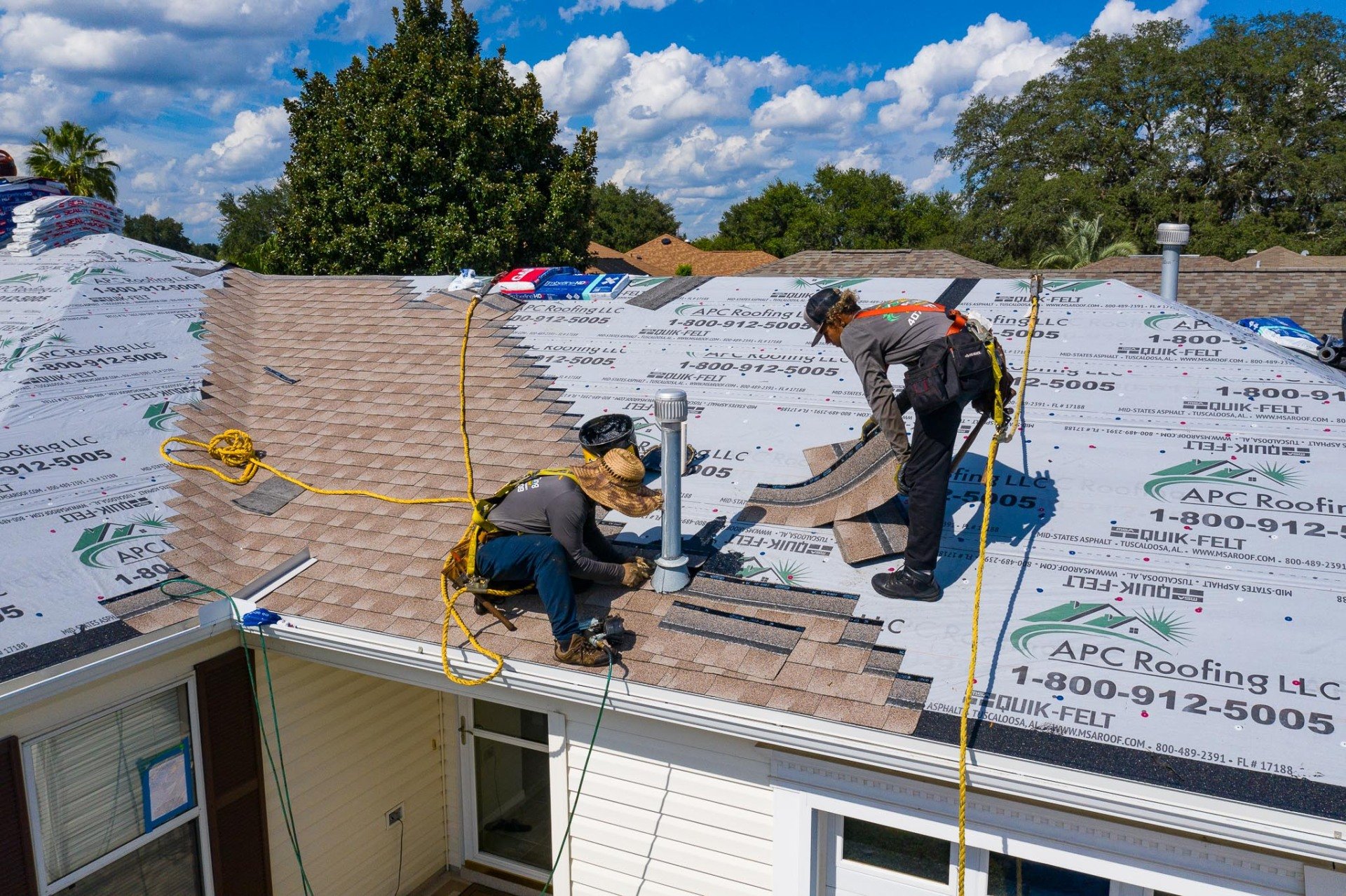 Two workers wearing safety harnesses install asphalt shingles on a house roof, surrounded by roofing underlayment and tools, on a sunny day. Two workers wearing safety harnesses install asphalt shingles on a house roof, surrounded by roofing underlayment and tools, on a sunny day.