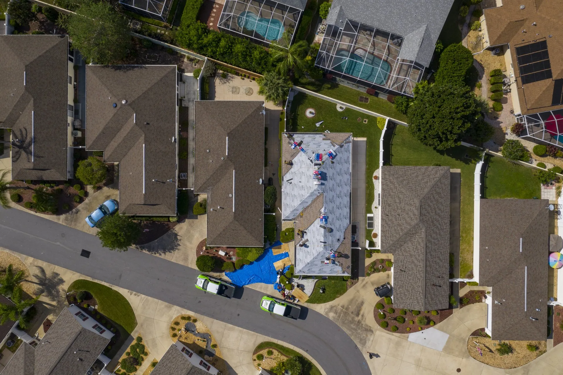 Aerial view of a residential neighborhood with workers installing a new roof on one house; two service vans and a blue tarp are visible on the driveway. Aerial view of a residential neighborhood with workers installing a new roof on one house; two service vans and a blue tarp are visible on the driveway.