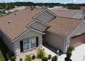 A beige single-story house with a brown shingle roof, two-car garage, neatly landscaped front yard, and adjacent neighboring homes.