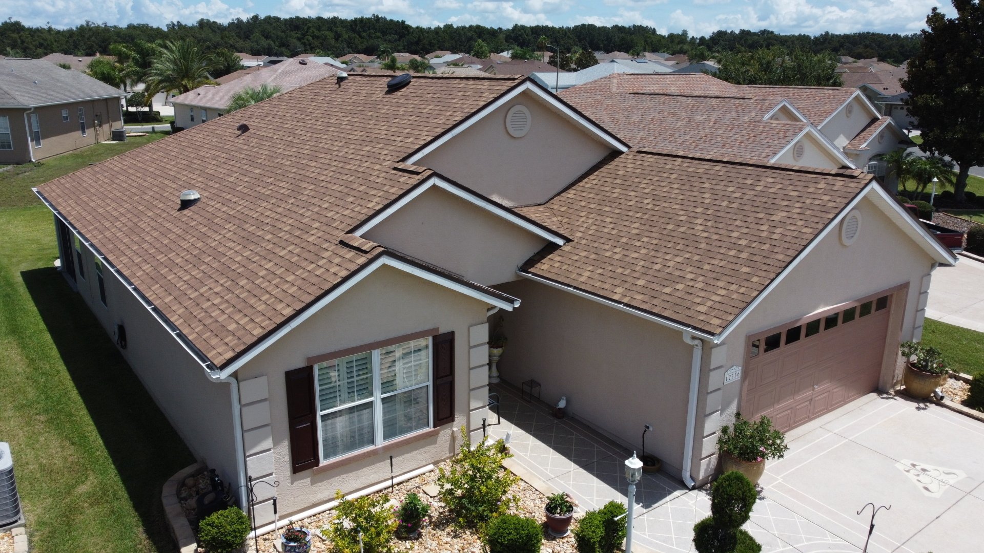 A beige single-story house with a brown shingle roof, two-car garage, neatly landscaped front yard, and adjacent neighboring homes.