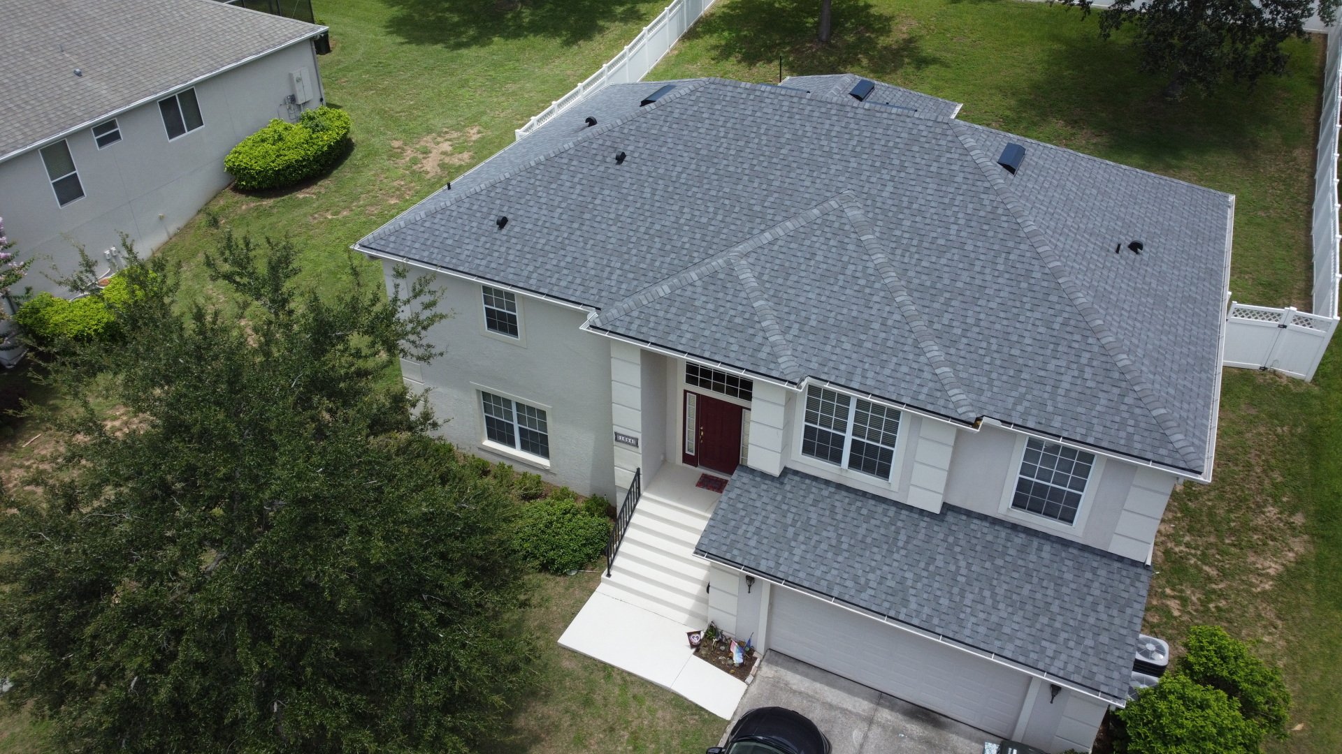 Aerial view of a two-story house with a gray shingle roof, white exterior, red front door, front steps, driveway, and surrounding green lawn.