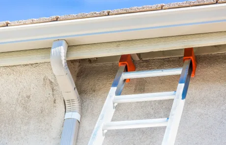 An aluminum ladder with orange stabilizers is propped against the roof gutter of a house, next to a white downspout, under a clear blue sky.