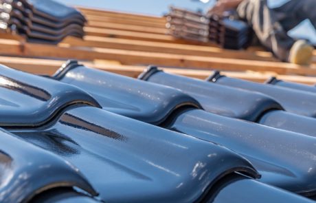 Close-up of glossy roof tiles being installed on a wooden roof frame, with a worker fitting tiles in the background under a clear sky.
