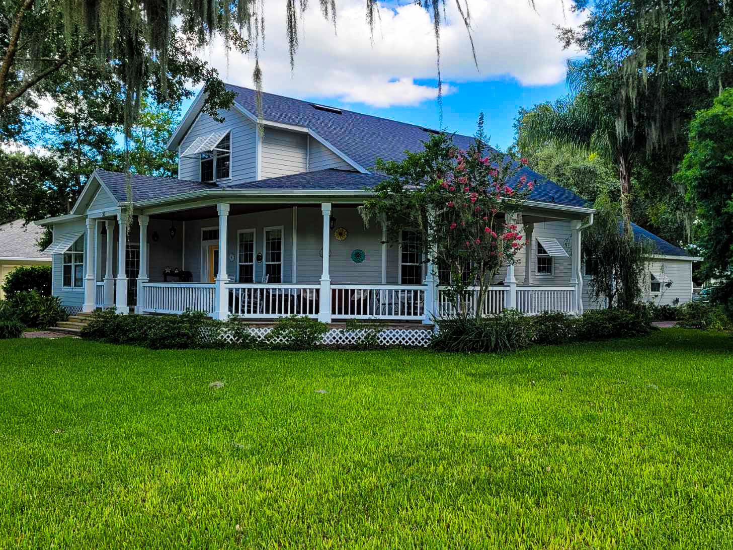 A white house with a large wraparound porch, surrounded by green grass, trees, and shrubs under a partly cloudy sky.