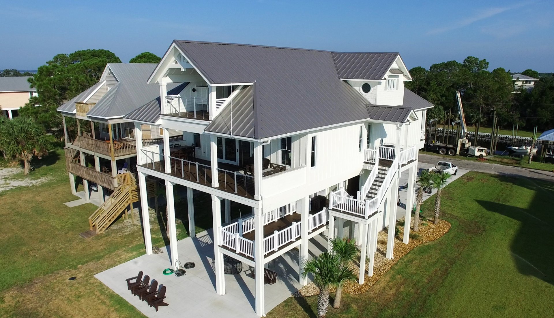 A large elevated white house with metal roof, multiple balconies, and a patio area, surrounded by grass and palm trees under a clear blue sky.