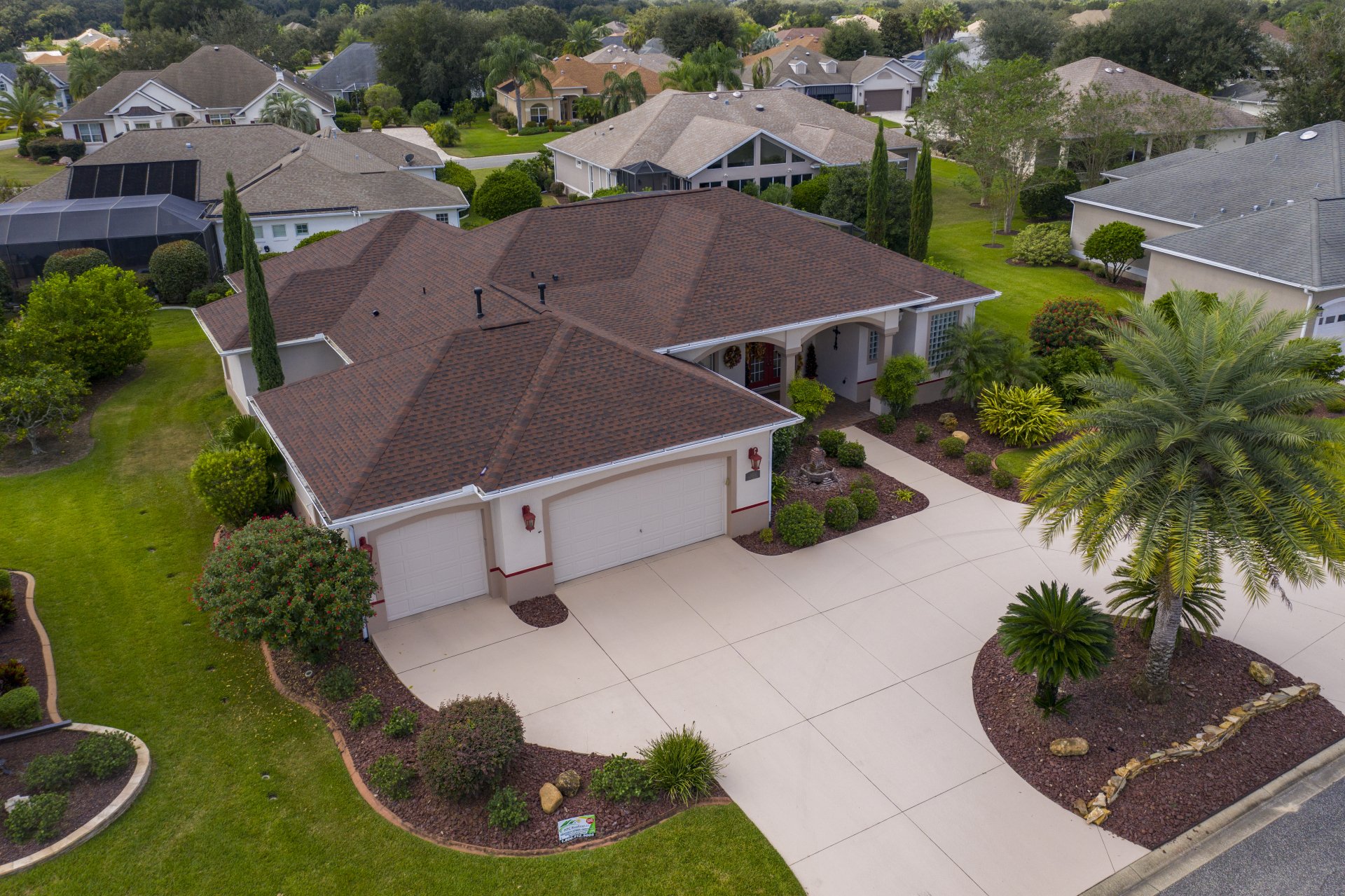 Aerial view of a single-story suburban house with a red roof, three-car garage, wide driveway, landscaped yard, and surrounding houses.