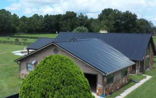 A large barn with a black metal roof sits on a grassy property surrounded by fenced pastures and trees, with horses visible in the background.