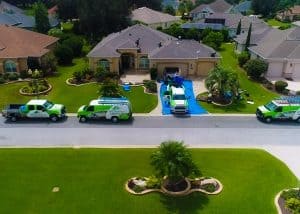Aerial view of four green and white service vehicles parked outside a suburban house where workers are performing maintenance on the roof and driveway.
