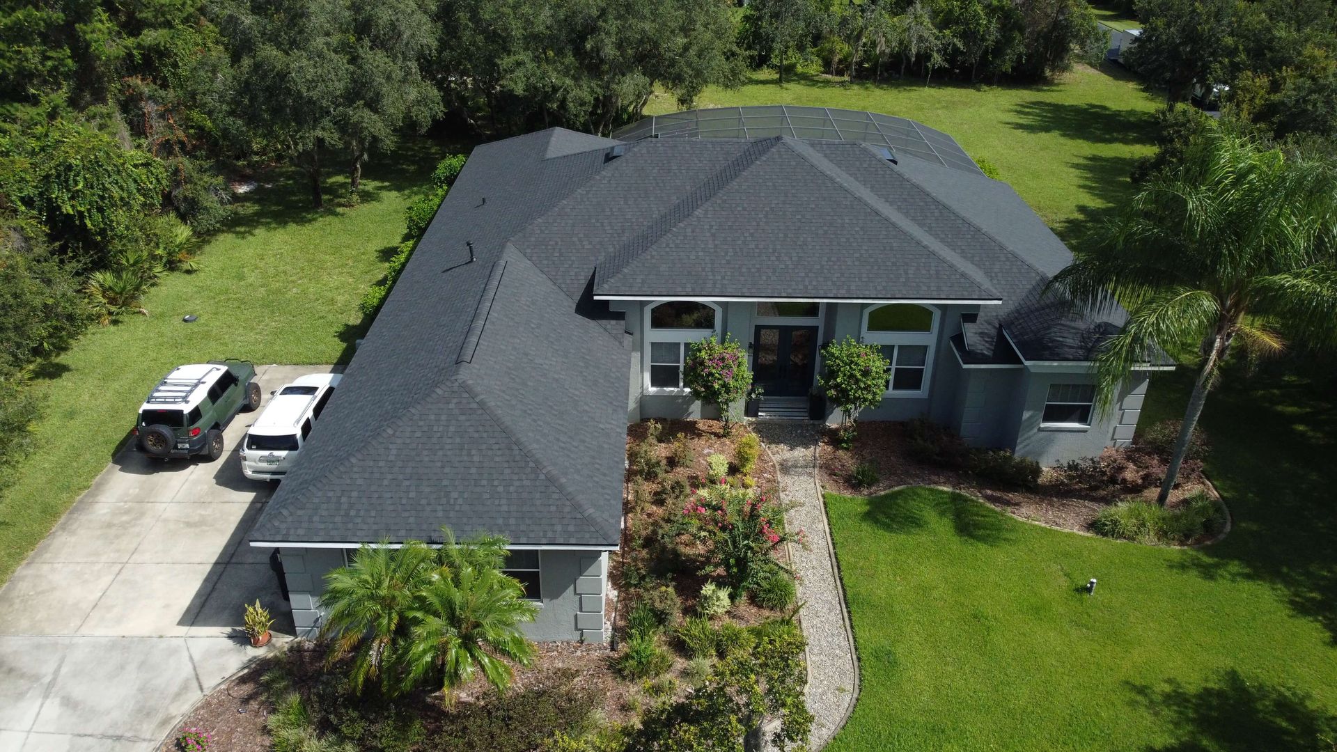 Aerial view of a large single-story house with a dark roof, two cars parked in the driveway, landscaped yard, and surrounding trees.
