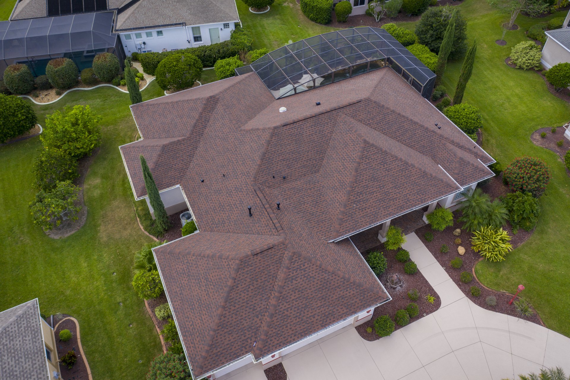 Aerial view of a house with a brown shingle roof, surrounded by landscaped lawns and shrubs, featuring a screened patio area at the back.
