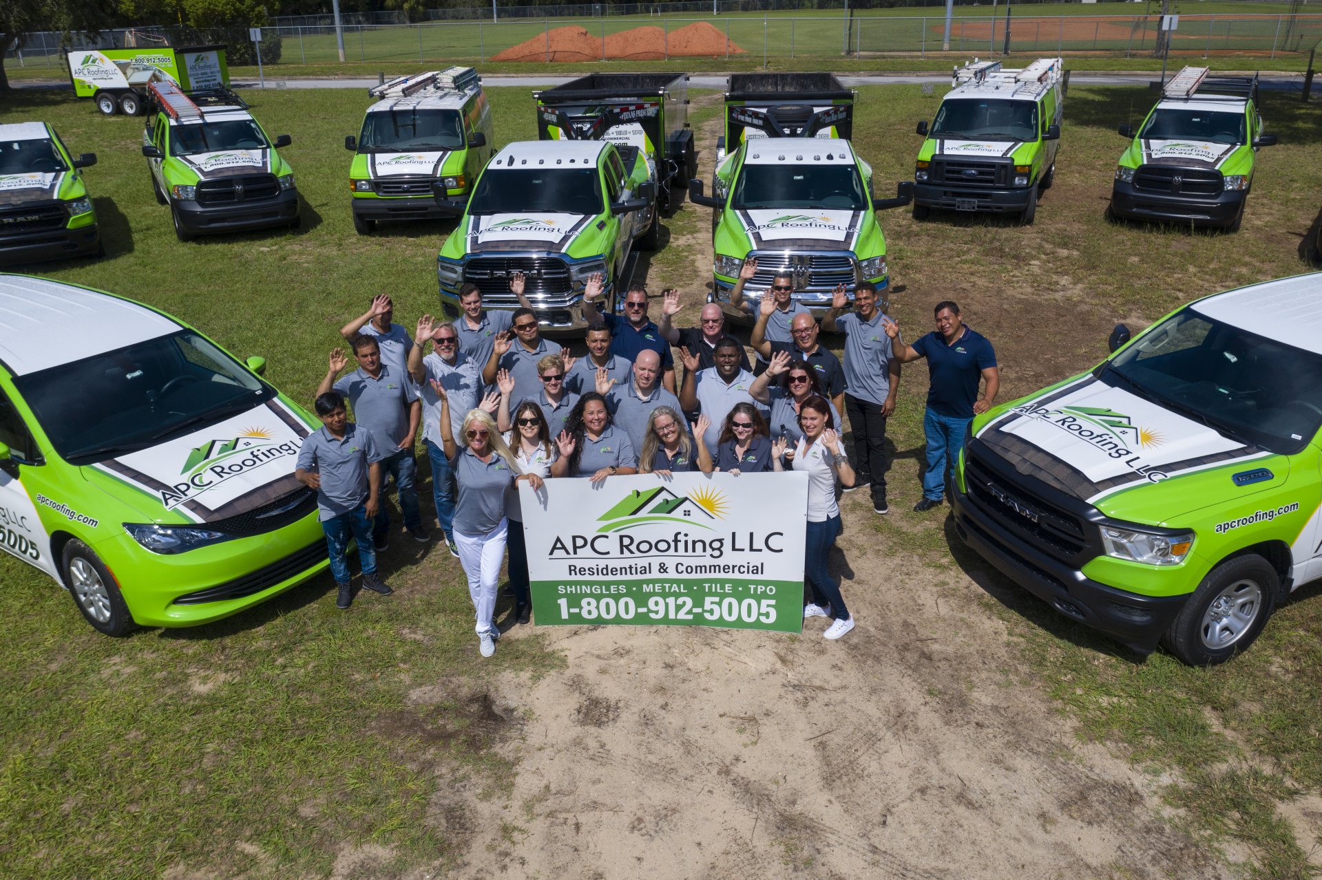 A group of people holding an APC Roofing LLC banner stands in front of several branded company trucks and vans in an outdoor area.