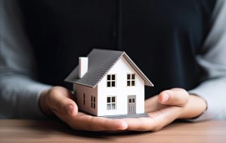 A person holding a small model house in their hands, with the house resting on their palms over a wooden surface.
