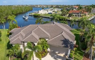 A large, modern house with a tiled roof sits near a canal with a docked sailboat, surrounded by palm trees and neighboring waterfront homes.