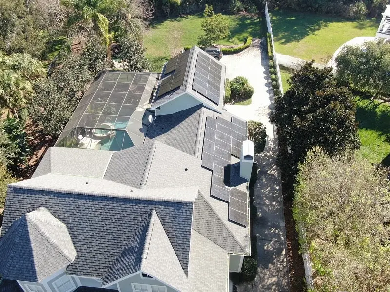 Aerial view of a house with multiple solar panels installed on its roof and a screened-in backyard pool area surrounded by trees.