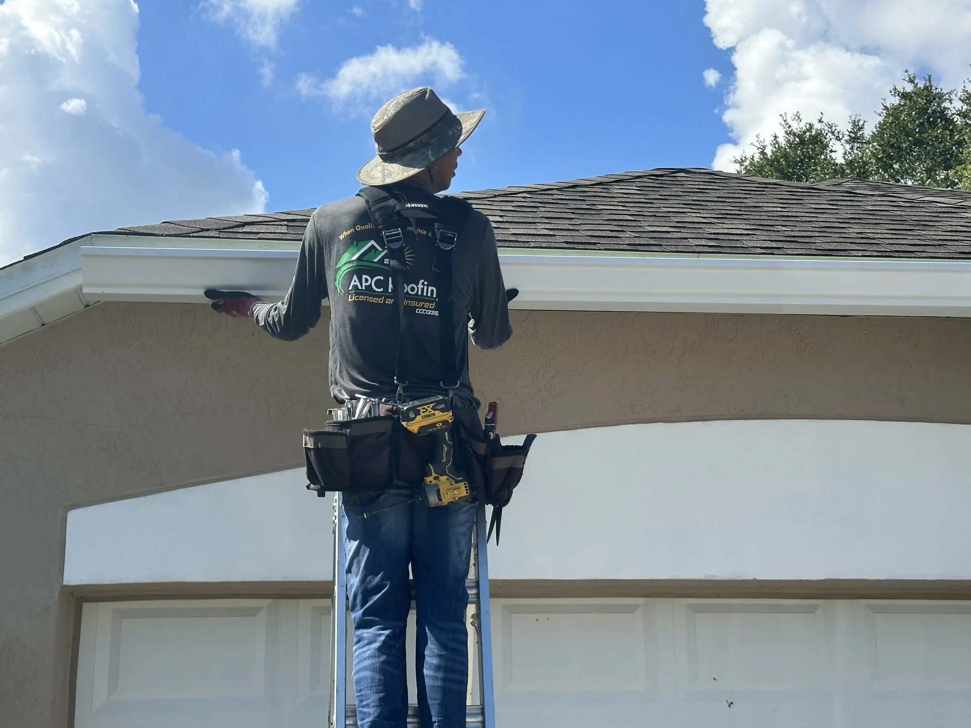 A worker on a ladder inspects or repairs the gutter of a house, wearing a tool belt and safety gear under a blue sky with clouds.
