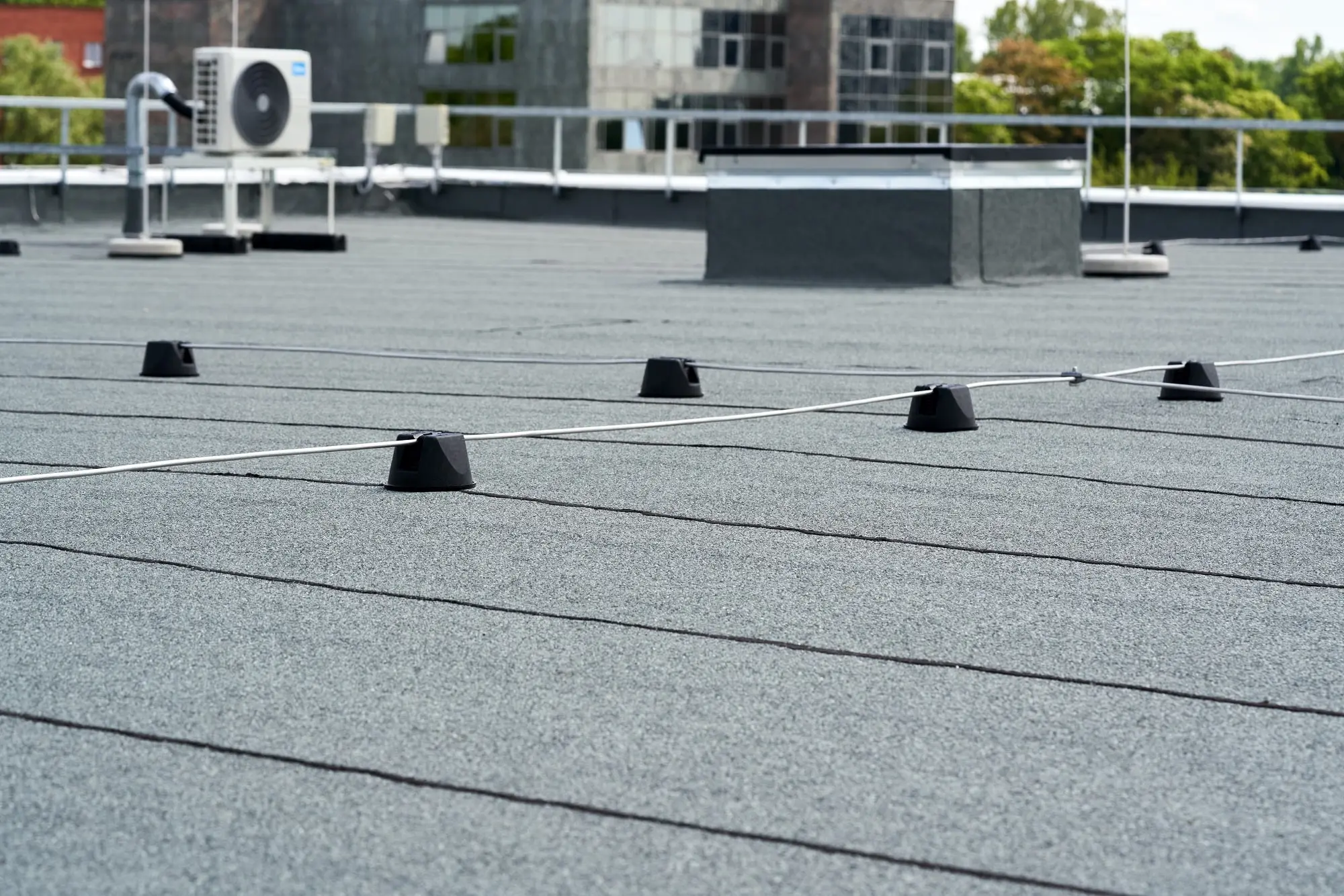 Flat commercial building roof with asphalt covering, safety wire system, and HVAC unit visible in the background.