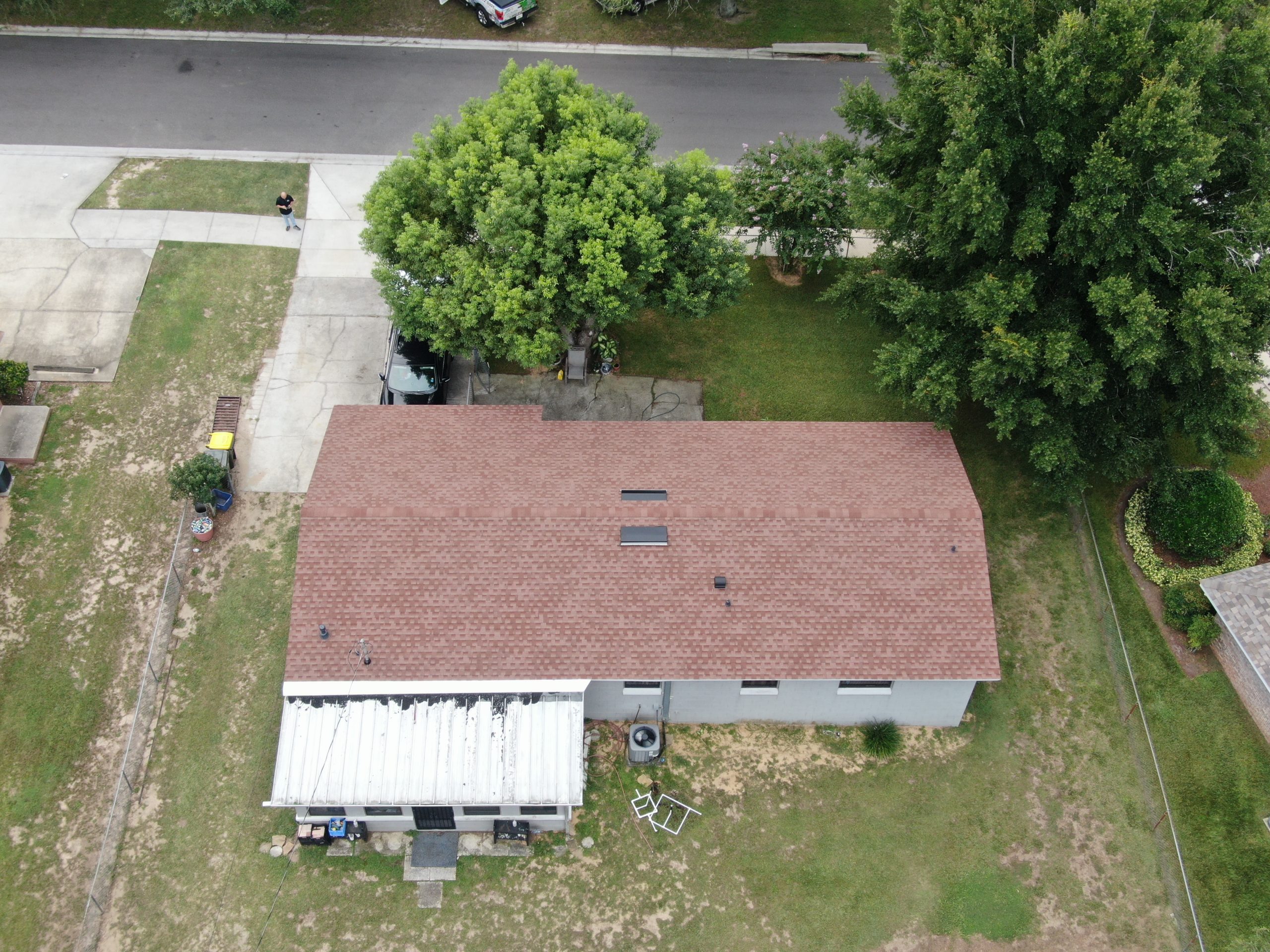 Aerial view of a single-story house with a brown roof, backyard, two large trees, and a partially covered patio. A driveway and street are visible at the top of the image.