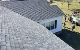 A view of a house roof with grey shingles, two people standing on the grass, and a trailer nearby on a sunny day.