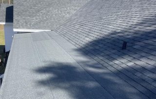 A house roof with gray asphalt shingles and an overhanging section; tree shadows are cast on the roof surface.