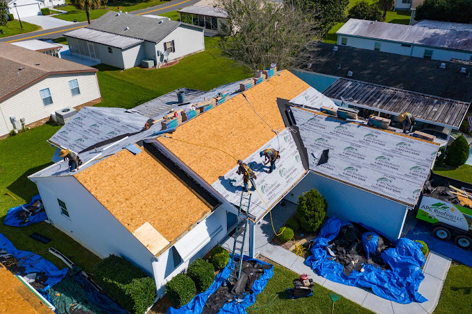 Aerial view of workers installing new roofing materials on a house, with tarps covering the ground and construction equipment nearby.