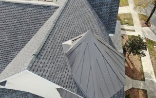 Aerial view of a building with gray asphalt shingle roofing and a metal roofed turret, surrounded by sidewalks, grass, and a few trees.