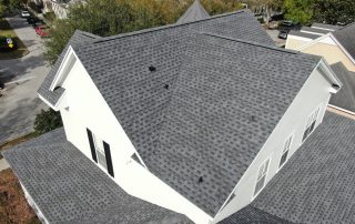 Aerial view of a residential house with a gray shingle roof, multiple roof sections, and white exterior walls in a suburban neighborhood.