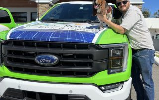 A man in sunglasses stands next to a green Ford truck with a dog on the hood, both outside a building. The truck advertises solar and gutter services.