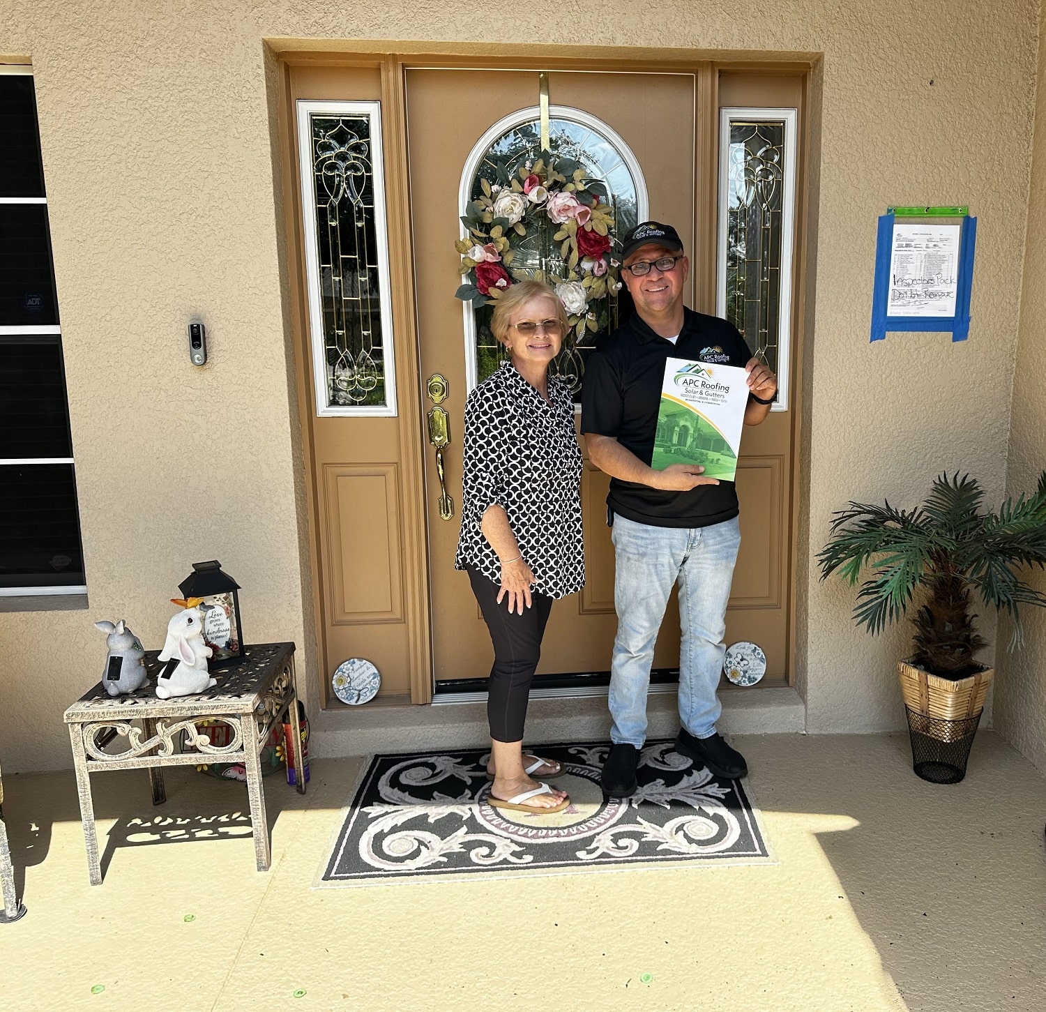 A satisfied homeowner and roofing representative pose in front of the home, proudly displaying the APC Roofing folder.