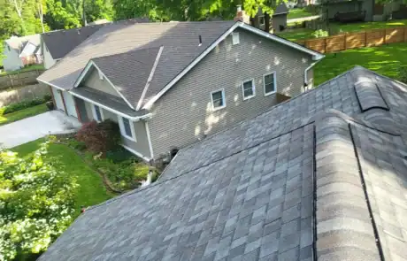 Residential roof with asphalt shingles and a steep pitch, surrounded by green trees and a backyard.